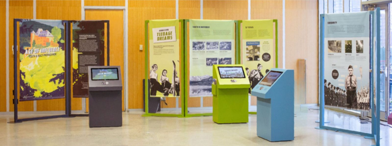 Age of Influence installed in a bright gallery: colourful freestanding panel units in green, purple and blue, each paired with a touchscreen kiosk